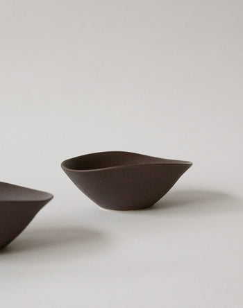 Minimalist photo of the NR Ceramics HIN Soup Bowl, dark and irregularly shaped, set on a plain light background with a partial glimpse of other versatile NR Ceramics tableware on the left edge.