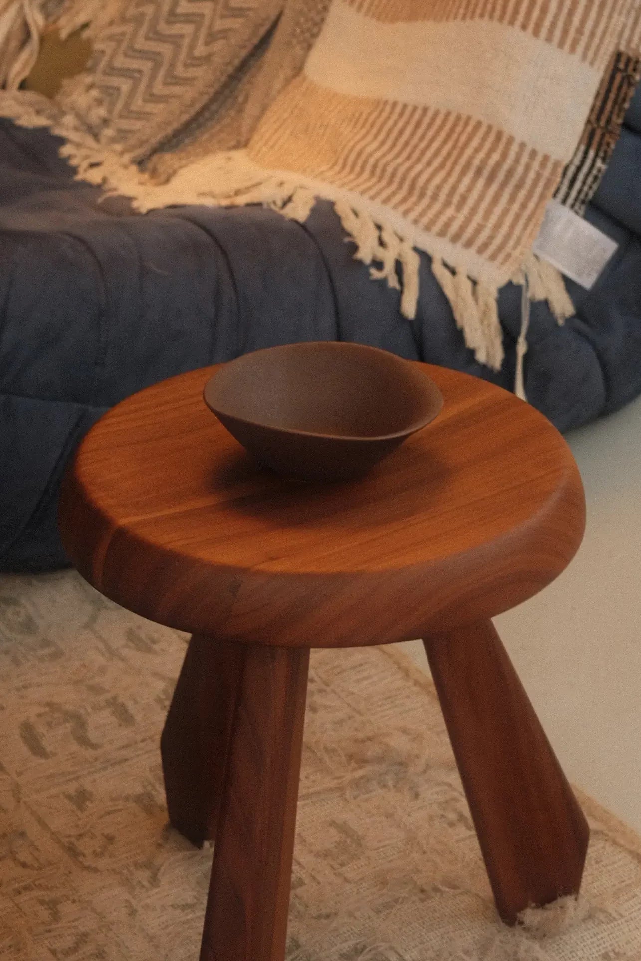 A small wooden side table with three legs holds an NR Ceramics HIN Soup Bowl. Behind it, a blue cushioned seat is draped with a beige striped tasseled blanket and patterned pillow, while a textured rug covers the floor.