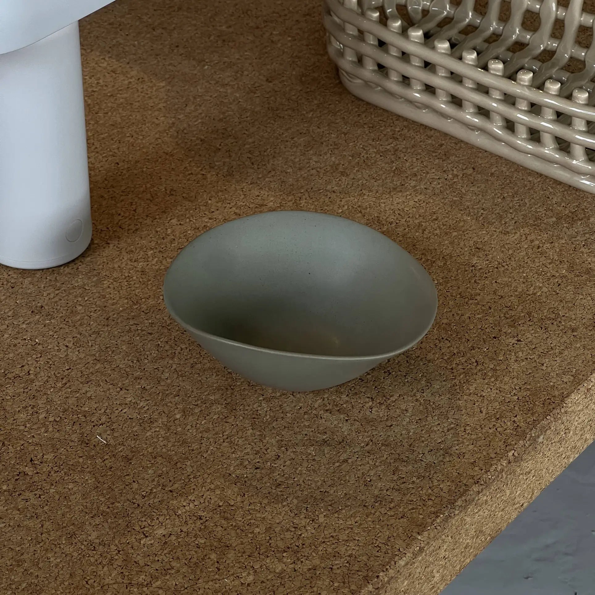The NR Ceramics HIN Soup Bowl, a small and smooth gray-green piece, sits on a cork surface beside a white cylindrical object and a beige woven basket, exemplifying versatile NR Ceramics tableware for any setting.