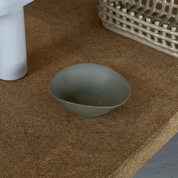 The NR Ceramics HIN Soup Bowl, a small and smooth gray-green piece, sits on a cork surface beside a white cylindrical object and a beige woven basket, exemplifying versatile NR Ceramics tableware for any setting.
