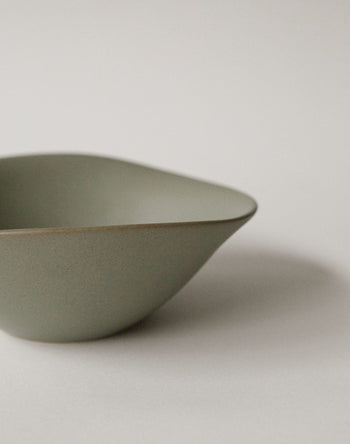The NR Ceramics HIN Soup Bowl is a light green, matte-finish piece with an asymmetrical modern shape. Partially visible against a white background, this minimalist tableware from NR Ceramics adds versatility to any setting.
