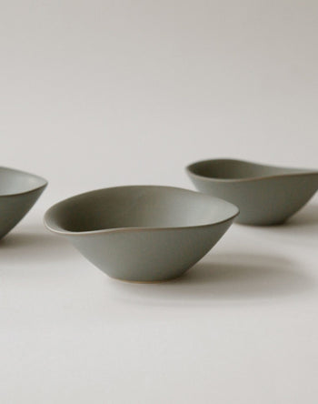 Three minimalist NR Ceramics HIN Soup Bowls in matte gray feature organic shapes. Spaced apart on a light background, these versatile bowls by NR Ceramics create a simple and elegant display.