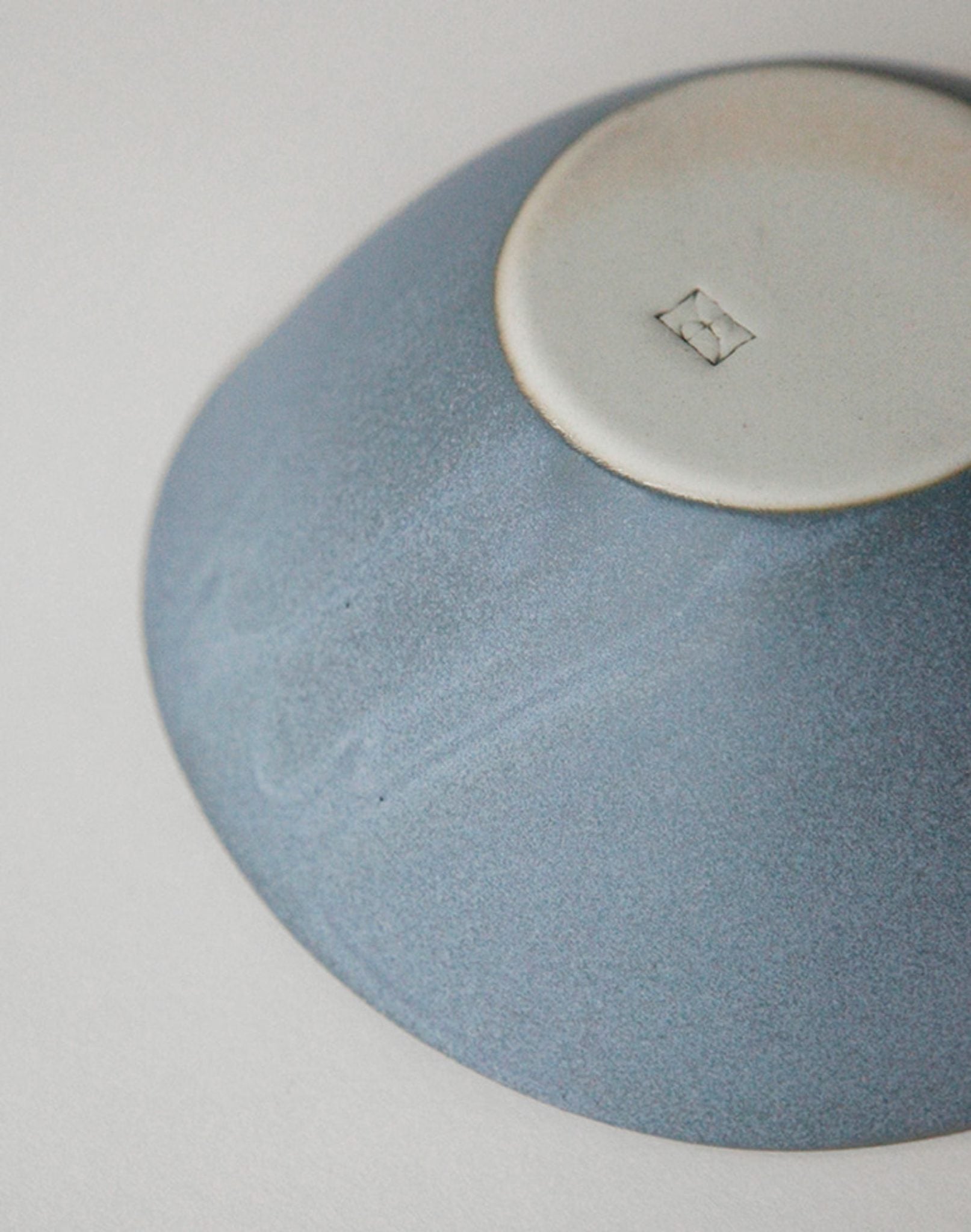 An NR Ceramics HIN Soup Bowl in blue ceramic rests upside down on a white surface, showing a small square makers mark on its unglazed base—versatile tableware by NR Ceramics for any setting.