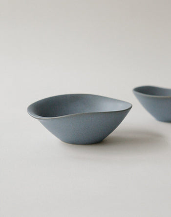 The NR Ceramics HIN Soup Bowl, a minimalist blue ceramic bowl with an organic, uneven rim, rests on a light surface. Another bowl from NR Ceramics versatile HIN collection is partially visible in the background.