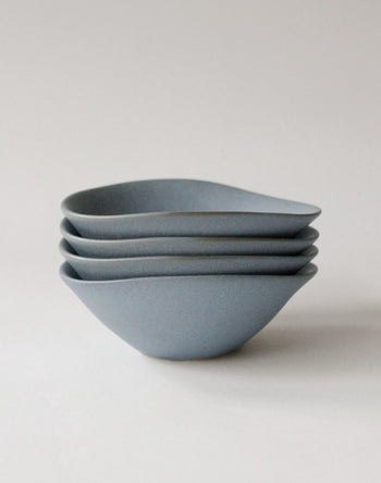 Four matte, light gray NR Ceramics HIN Soup Bowls, each with a subtly organic shape, are stacked on a plain background—versatile tableware from NR Ceramics for any occasion.