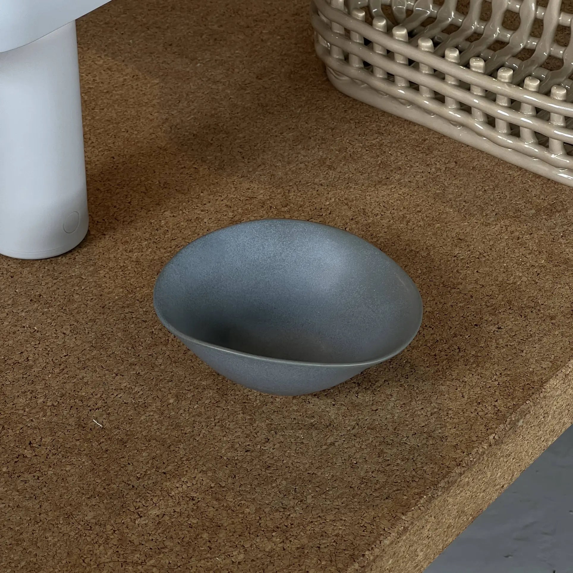 The NR Ceramics HIN Soup Bowl, a small matte gray piece from NR Ceramics, rests on a cork surface near a white cylindrical item and beige woven basket, highlighting its versatile design for any table setting.