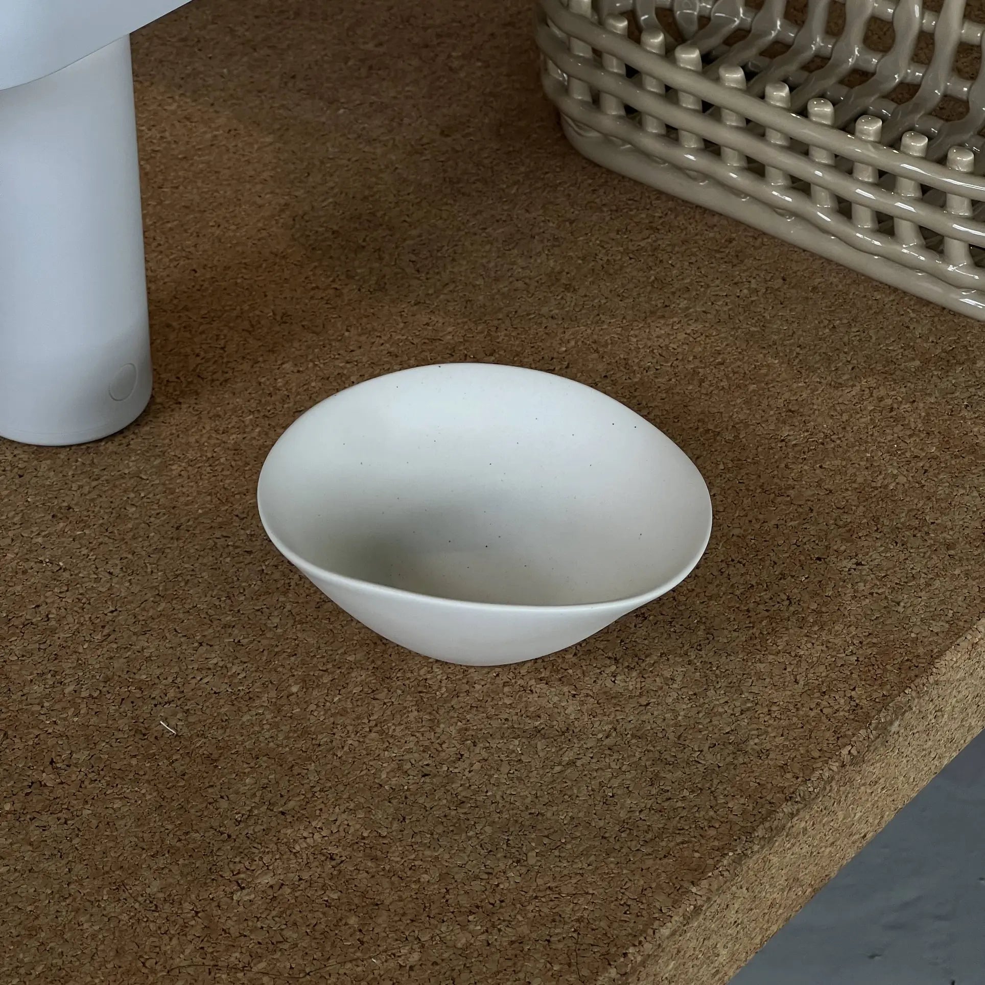 The NR Ceramics HIN Soup Bowl, a small white ceramic piece by NR Ceramics, rests on a cork surface beside a white cylinder and a woven beige basket.