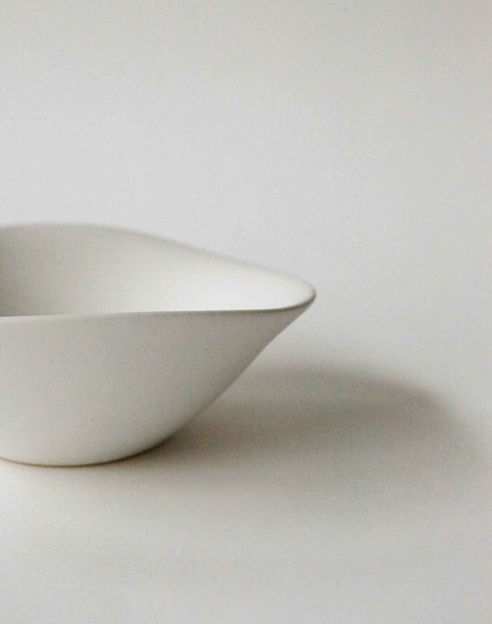 A minimalist white NR Ceramics HIN Soup Bowl with smooth curves sits on a plain surface, photographed against a light background. Only part of the bowl is visible, highlighting its simple, modern design by NR Ceramics.