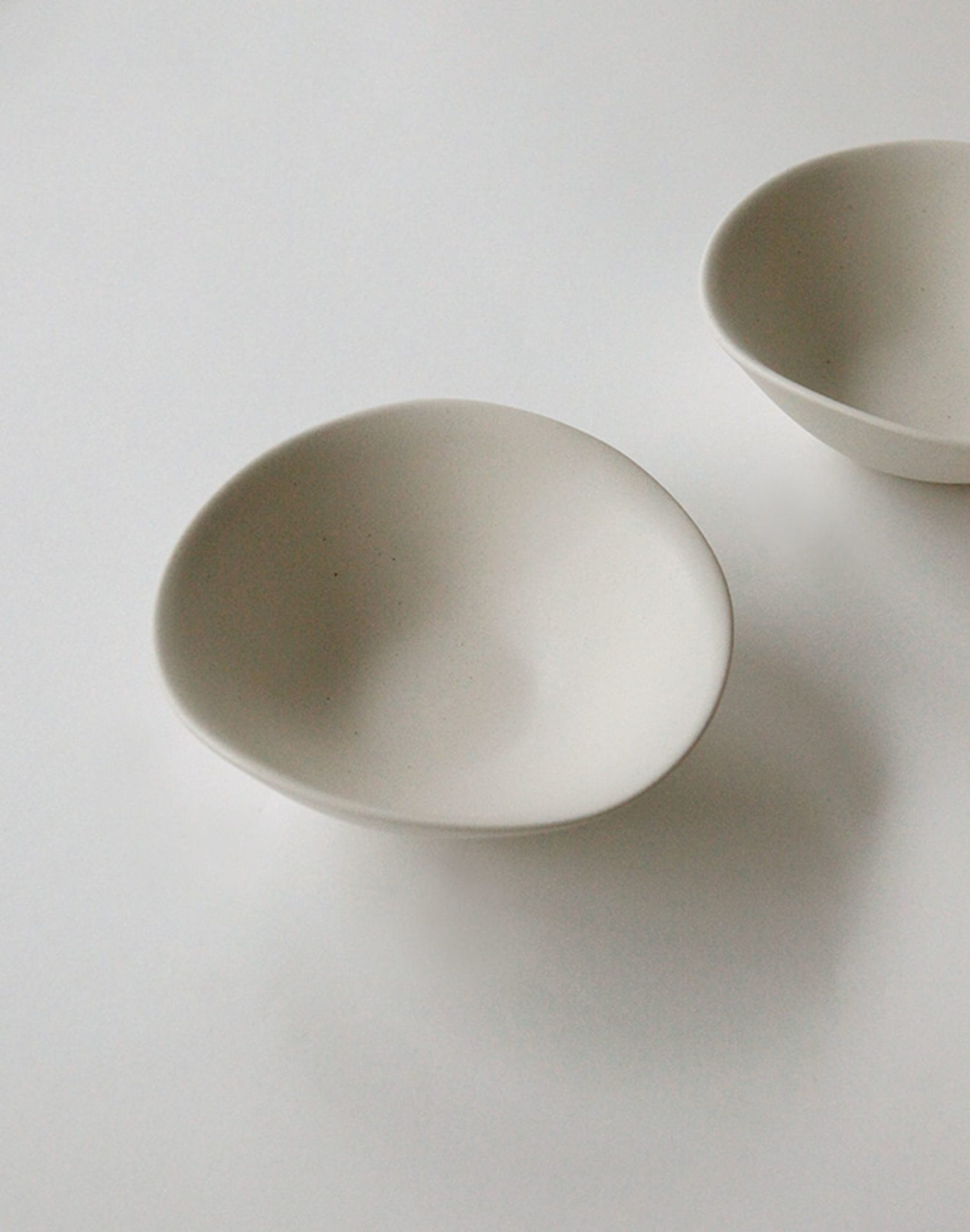 Two minimalist white NR Ceramics HIN Soup Bowls by NR Ceramics sit on a plain white surface; one bowl is centered in front, while the other versatile piece is partially shown in the upper right corner.