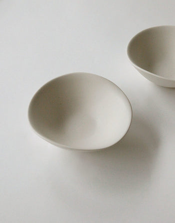 Two minimalist white NR Ceramics HIN Soup Bowls by NR Ceramics sit on a plain white surface; one bowl is centered in front, while the other versatile piece is partially shown in the upper right corner.