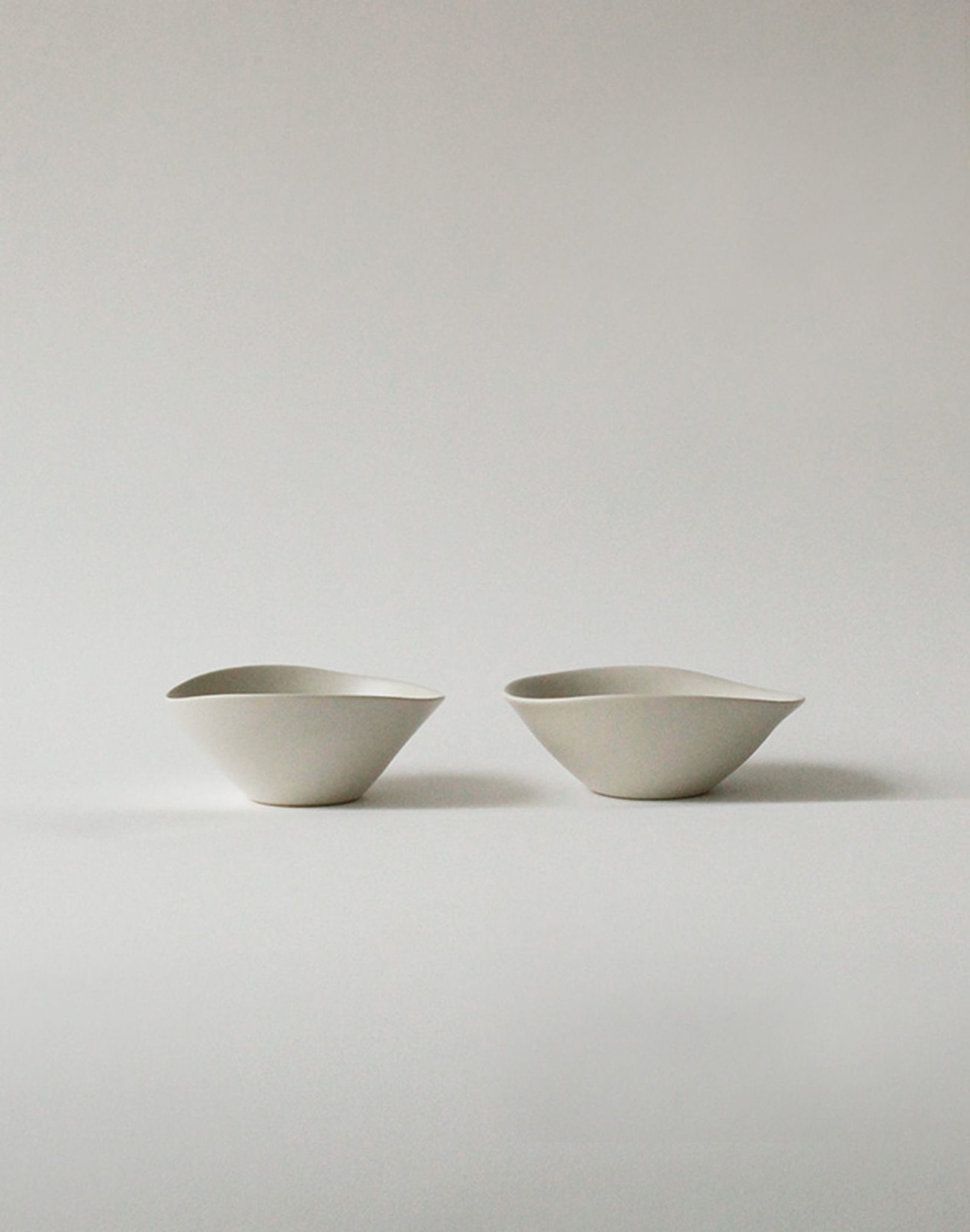 Two minimalist, asymmetrical NR Ceramics HIN Soup Bowls are set side by side on a plain, light gray surface. With their modern abstract design, these versatile bowls by NR Ceramics are ideal for soup or serving a variety of dishes.
