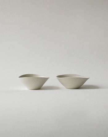 Two minimalist, asymmetrical NR Ceramics HIN Soup Bowls are set side by side on a plain, light gray surface. With their modern abstract design, these versatile bowls by NR Ceramics are ideal for soup or serving a variety of dishes.