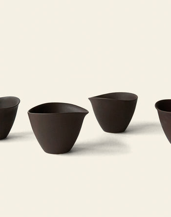 Four dark brown NR Ceramics HIN Tea Cups with a minimalist, ergonomic design and pointed lips are arranged on a light background, casting soft shadows for a clean, modern look.