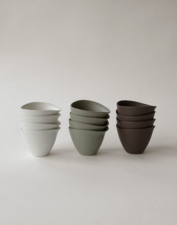 Three stacks of NR Ceramics HIN Tea Cups in white, muted green, and dark brown are arranged side by side against a plain light background. Each stack from NR Ceramics includes three ergonomically designed HIN Tea Cups.