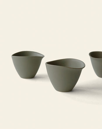 Three minimalist, matte olive green NR Ceramics HIN Tea Cups with ergonomic, organic shapes are arranged on a plain off-white background. Only part of the third cup on the right is visible.