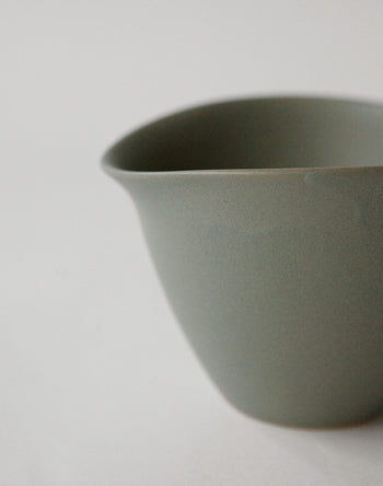 A close-up of the NR Ceramics HIN Tea Cup by NR Ceramics, featuring a simple matte grey finish, smooth surface, and slightly curved rim. The ergonomic design is highlighted against a light background, with the cup partially cropped on the right.