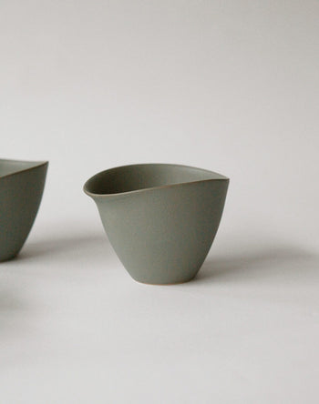 A minimalist gray NR Ceramics HIN Tea Cup with an asymmetrical wavy rim and ergonomic design rests on a white surface, while another NR Ceramics HIN cup is partially visible on the left.