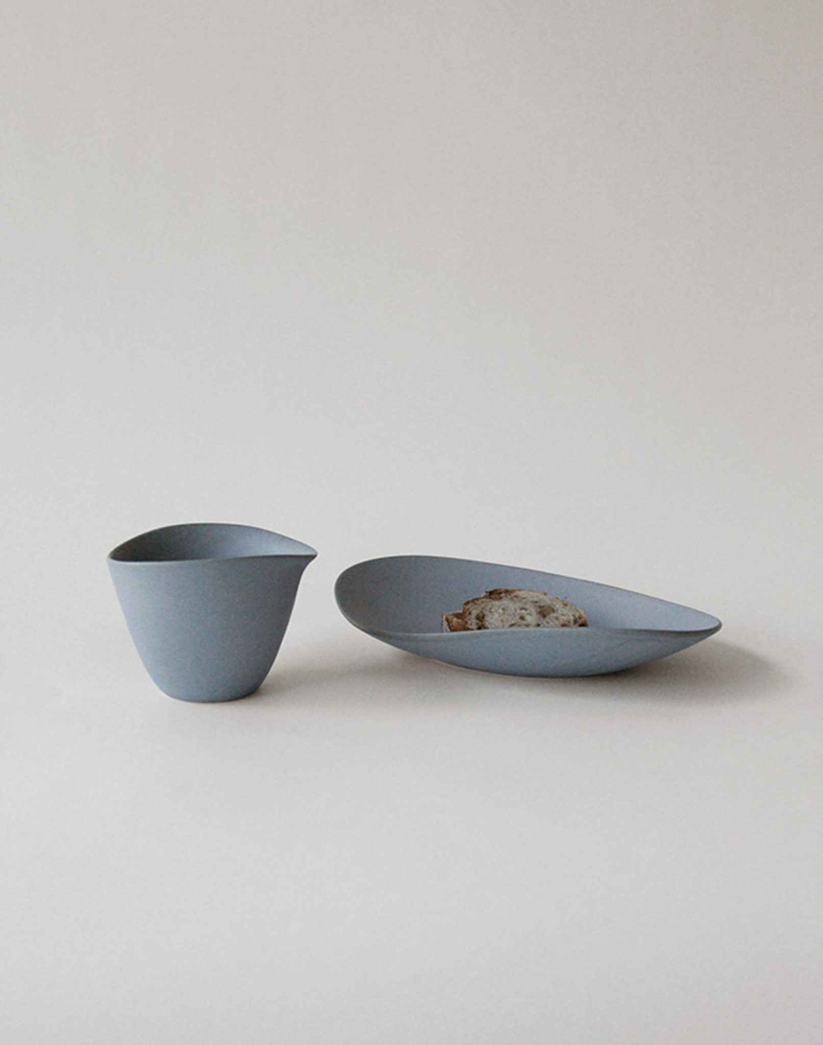 A minimalist scene with the NR Ceramics HIN Tea Cup, known for its ergonomic design, and a matching oval plate holding a slice of bread, set against a plain light background.