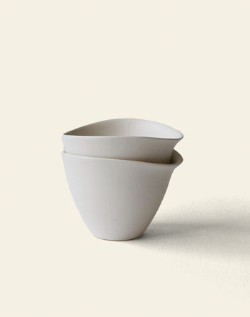 Two minimalist, off-white NR Ceramics HIN Tea Cups are stacked on a light background. Their simple, slightly asymmetrical shape highlights ergonomic design for comfortable daily use.