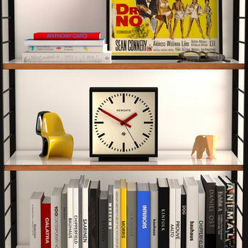 A bookshelf displays a Newgate Amp Mantel Clock, books, a yellow abstract sculpture, and an elephant figurine. The top shelf features a vintage Dr. No movie poster and an Anthony Caro book for modern style.