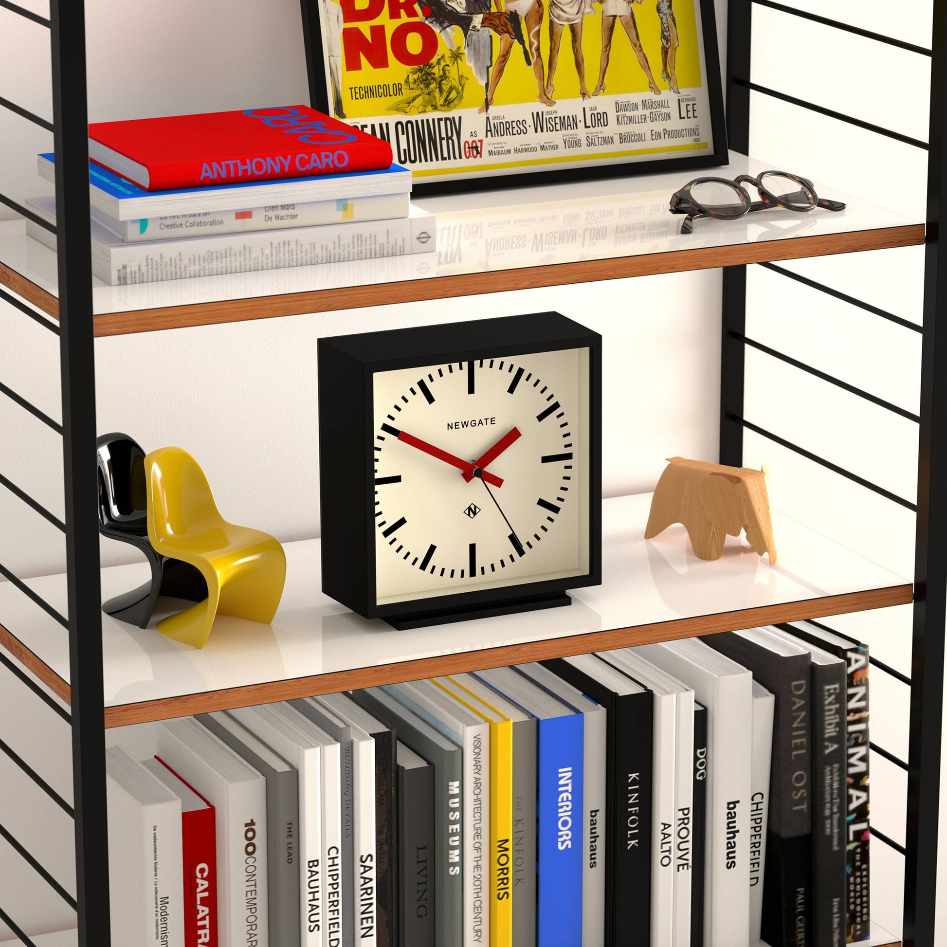 A bookshelf with books, a Newgate Amp Mantel Clock in black and white, a small yellow and black chair model, a wooden decorative object, plus an upper shelf with magazines and a small red car model.