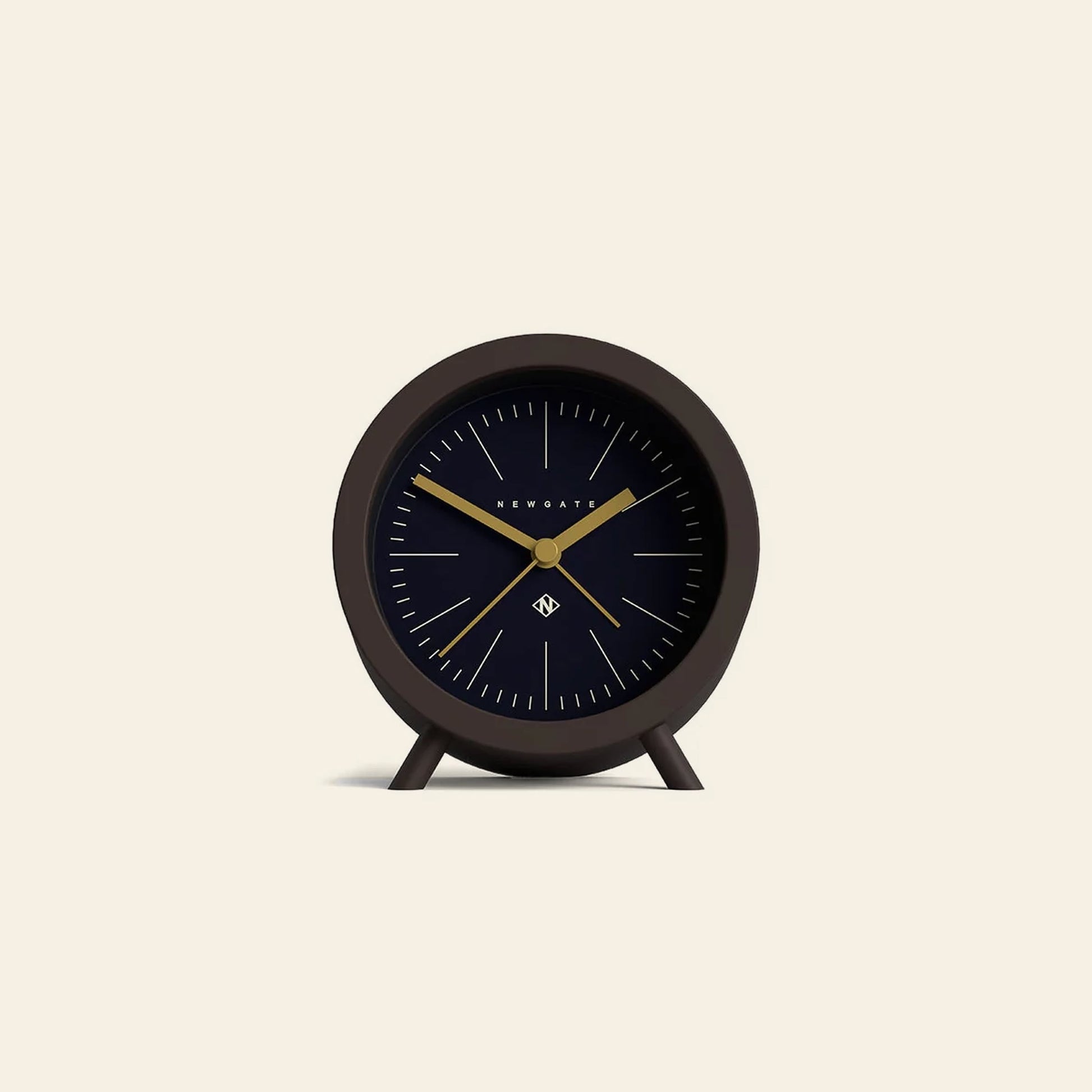 The Newgate Fred Alarm Clock is a minimalist black analog clock with gold hands and simple hour marks. Ideal for a mid-century modern bedside table, it stands on two small legs against a light beige background. Brand: Newgate.