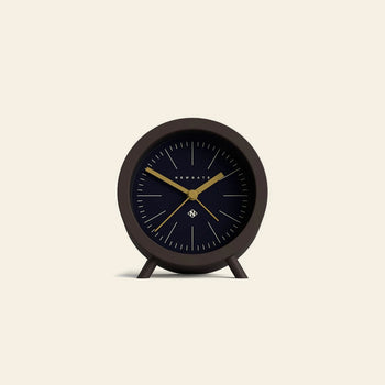 The Newgate Fred Alarm Clock is a minimalist black analog clock with gold hands and simple hour marks. Ideal for a mid-century modern bedside table, it stands on two small legs against a light beige background. Brand: Newgate.