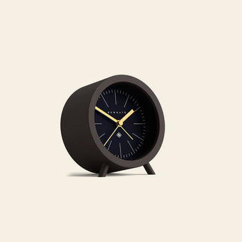 The Newgate Fred Alarm Clock by Newgate features a modern black design with gold hands and minimalist white markers, inspired by mid-century style. It stands on two legs—ideal for any bedside table against a plain off-white background.