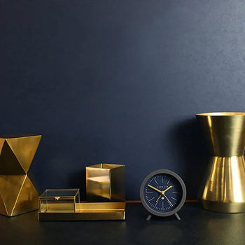 A set of gold and brass desk accessories, including a geometric sculpture, box, pencil holder, and vase, sits next to the Newgate Fred Alarm Clock by Newgate with gold hands against a dark blue background.