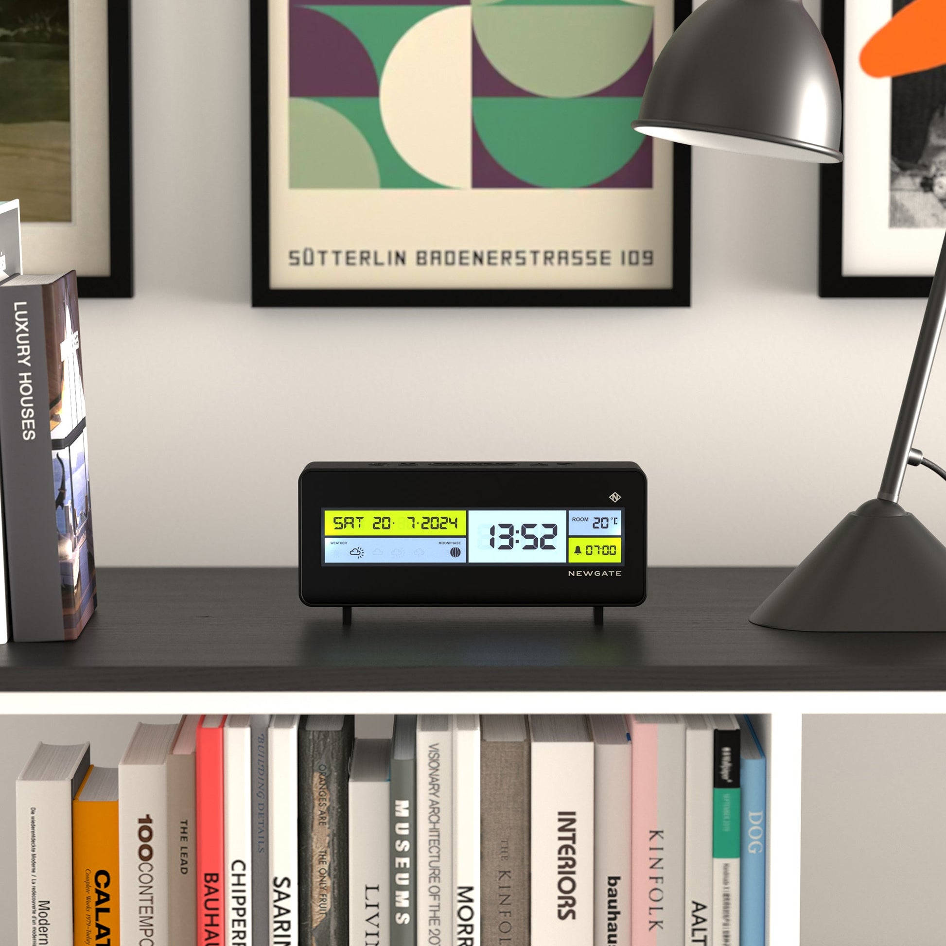The Newgate Futurama LCD Alarm Clock by Newgate sits on a shelf beside a desk lamp, framed abstract art, and books. Its display shows the date, time, temperature, and alarm settings.