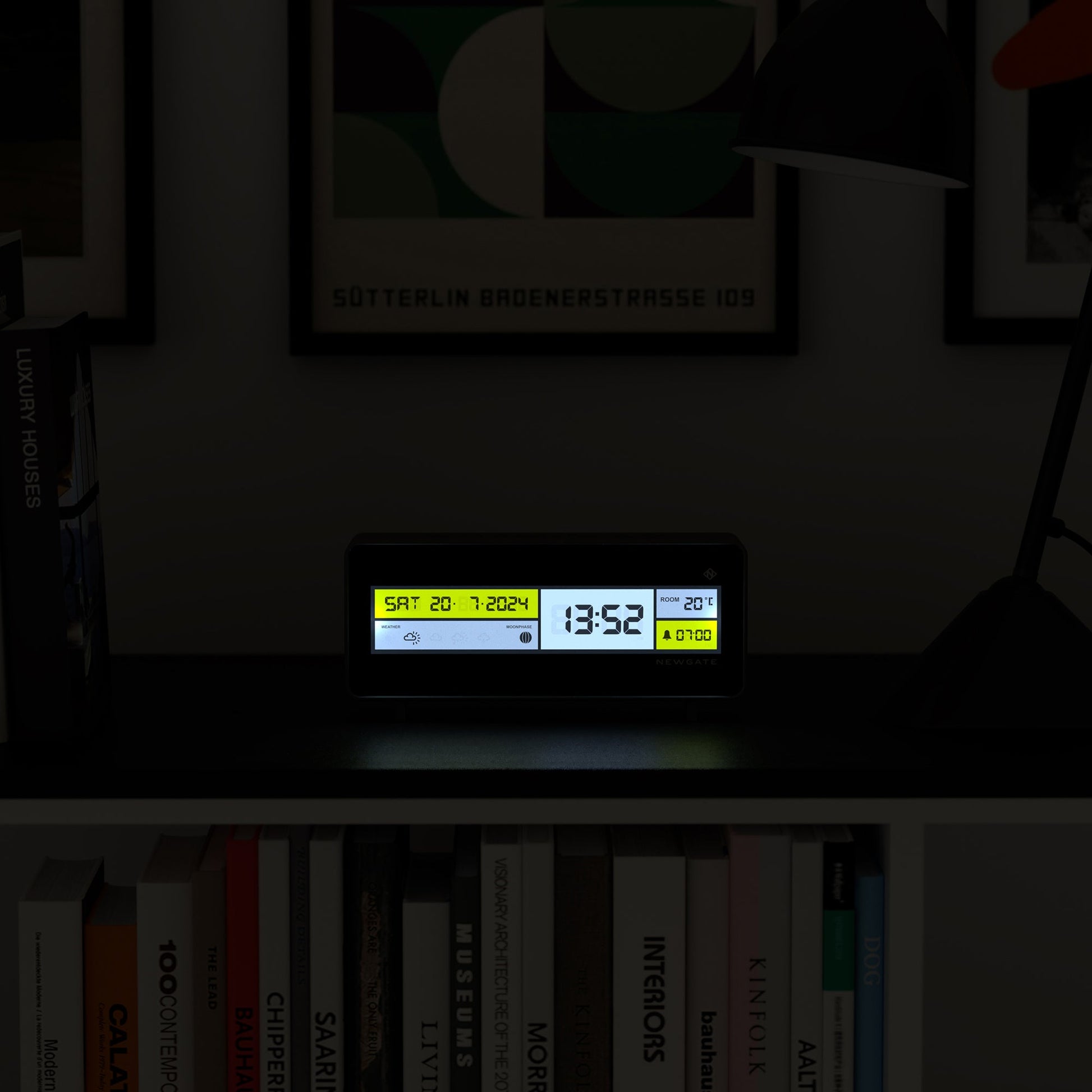The Newgate Futurama LCD Alarm Clock by Newgate sits on a dark shelf, displaying Sat 20-7-2024, 13:52, and 20°C. A yellow alarm icon glows as books and framed wall art decorate the background.