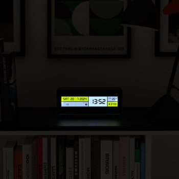 The Newgate Futurama LCD Alarm Clock by Newgate sits on a dark shelf, displaying Sat 20-7-2024, 13:52, and 20°C. A yellow alarm icon glows as books and framed wall art decorate the background.