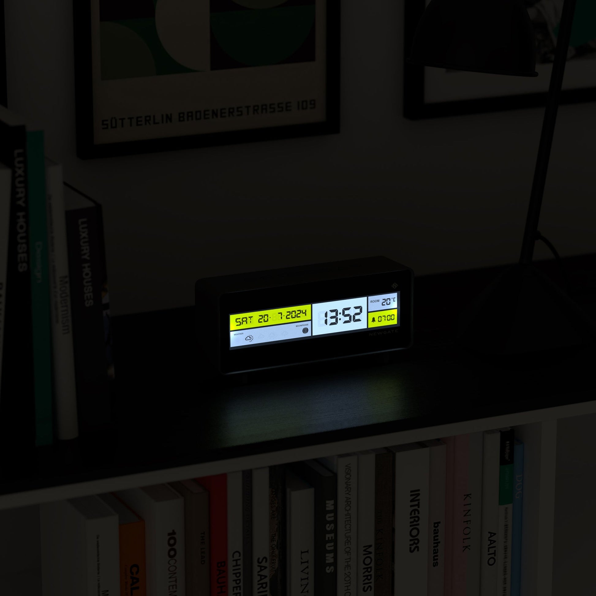 The Newgate Futurama LCD Alarm Clock by Newgate displays date, time, and temperature on a dark shelf, surrounded by books and a desk lamp in a dimly lit room.