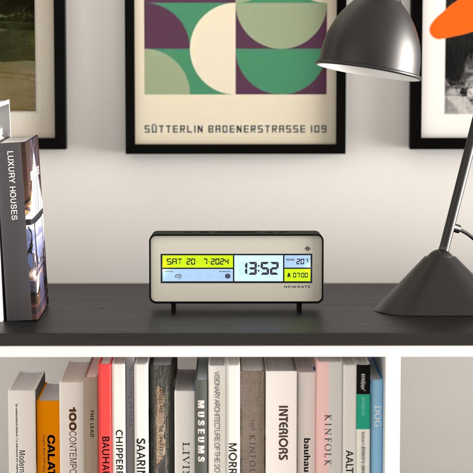 A Newgate Futurama LCD Alarm Clock by Newgate sits on a dark shelf, showing the date Sat 20-7-2024 and time 13:52. Books, a lamp, and framed art decorate the background.