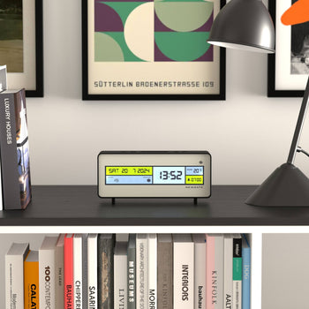 A Newgate Futurama LCD Alarm Clock by Newgate sits on a dark shelf, showing the date Sat 20-7-2024 and time 13:52. Books, a lamp, and framed art decorate the background.