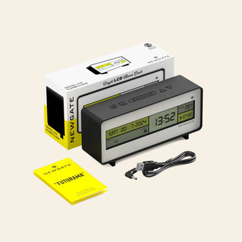 The Newgate Futurama LCD Alarm Clock with dual screens and temperature display is shown beside its packaging, a yellow instruction manual, and a USB charging cable, all set against a plain light background.