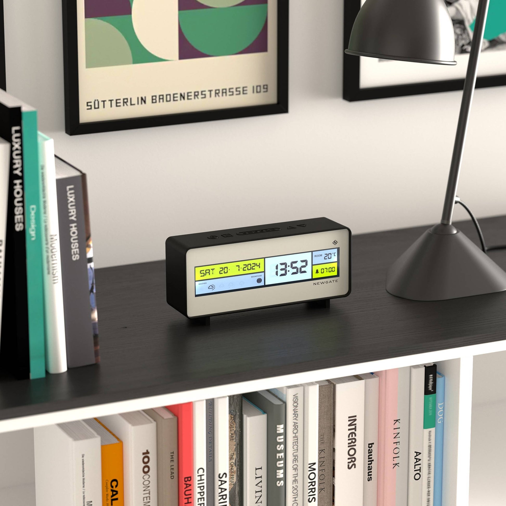 The Newgate Futurama LCD Alarm Clock by Newgate, showing time, date, and temperature on its screen, sits on a black shelf near a lamp, with books and framed artwork in the background.