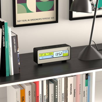 The Newgate Futurama LCD Alarm Clock by Newgate, showing time, date, and temperature on its screen, sits on a black shelf near a lamp, with books and framed artwork in the background.