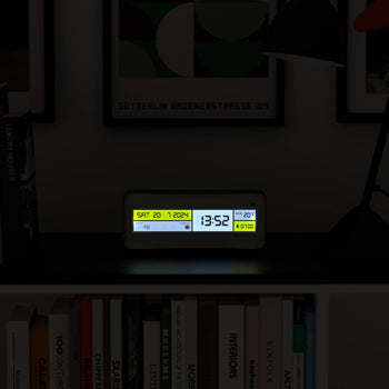 The Newgate Futurama LCD Alarm Clock by Newgate sits on a dark shelf, displaying SAT 20-7-2024 and 13:52. Books and framed artwork can be seen in the background.