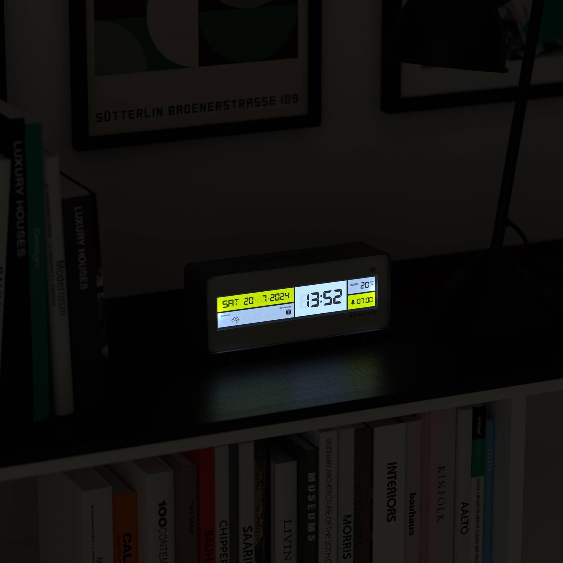 The Newgate Futurama LCD Alarm Clock by Newgate displays the date, time, and temperature with a bright LCD screen, illuminating a dark shelf surrounded by books and framed artwork in the background.