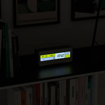 The Newgate Futurama LCD Alarm Clock by Newgate displays the date, time, and temperature with a bright LCD screen, illuminating a dark shelf surrounded by books and framed artwork in the background.