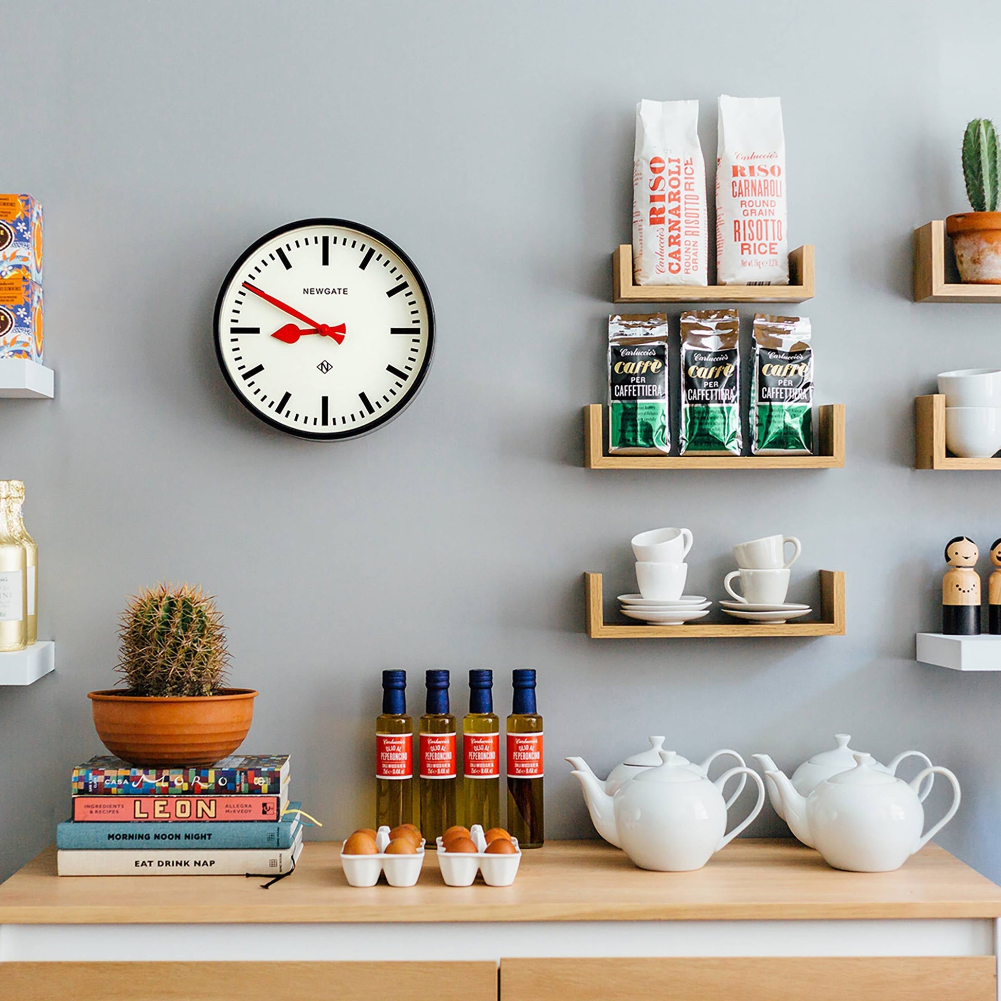 A modern kitchen wall featuring the Newgate Luggage Wall Clock by Newgate brings retro style above shelves with cups, jars, coffee, risotto rice, and teapots. Cookbooks, eggs, a potted cactus, and bottles line the wooden countertop.