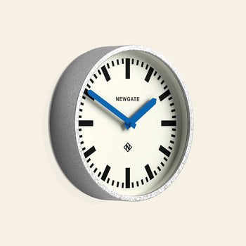 The Newgate Luggage Wall Clock Galvanised features a gray frame, white face, black markers, and blue hands. Inspired by mid-century railway station clocks, it displays the Newgate branding on its face.