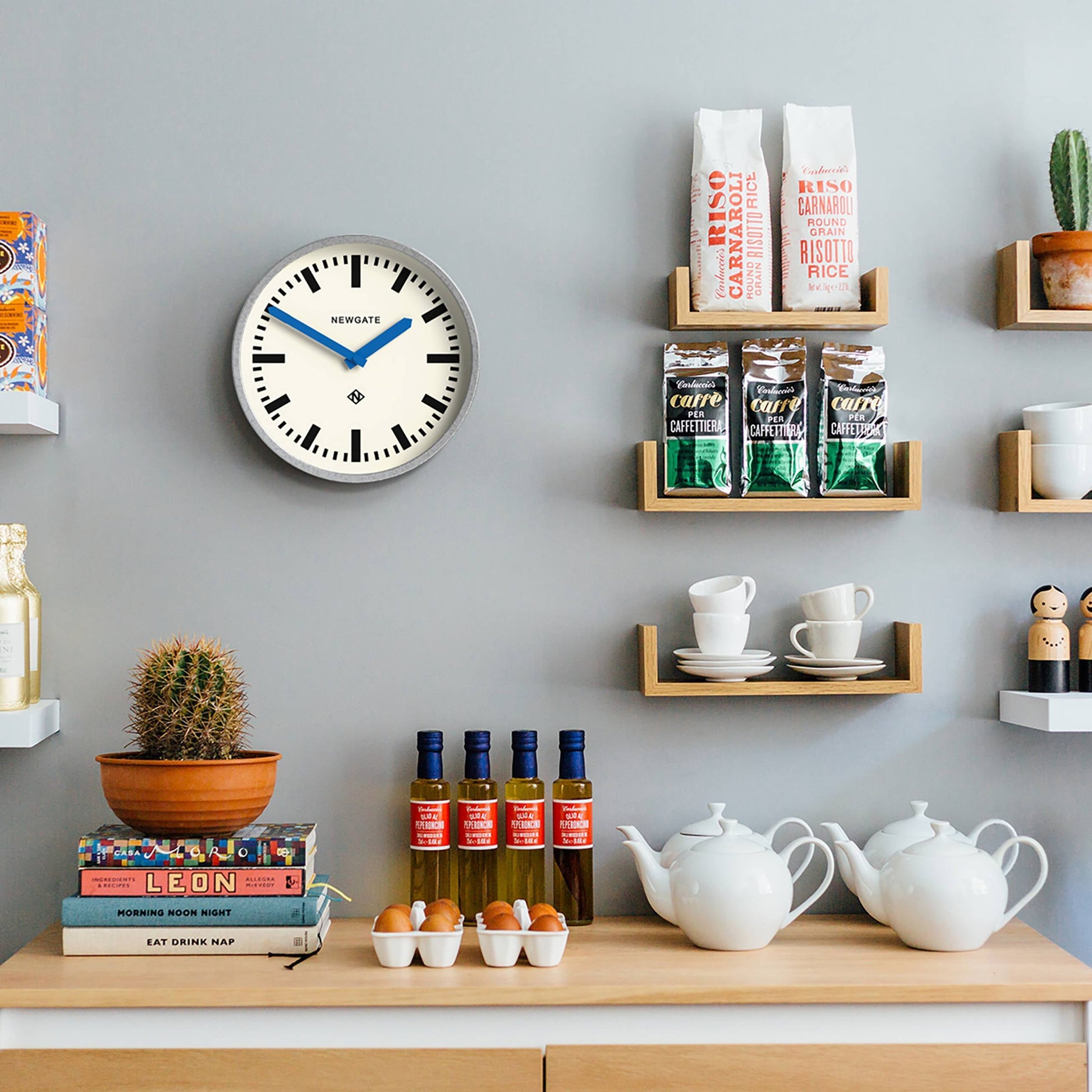 A kitchen wall with shelves holding coffee, rice, cups, teapots, sauces, eggs, a cactus, books, and décor. A Newgate Luggage Wall Clock Galvanised by Newgate shows 10:10 against a light gray background.