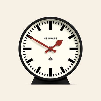 The Newgate M Mantel Railway Clock features a classic round black frame, bold hour marks, red hands set to 10:09:39, and stands on a small black base with NEWGATE displayed at the top of the clock face.