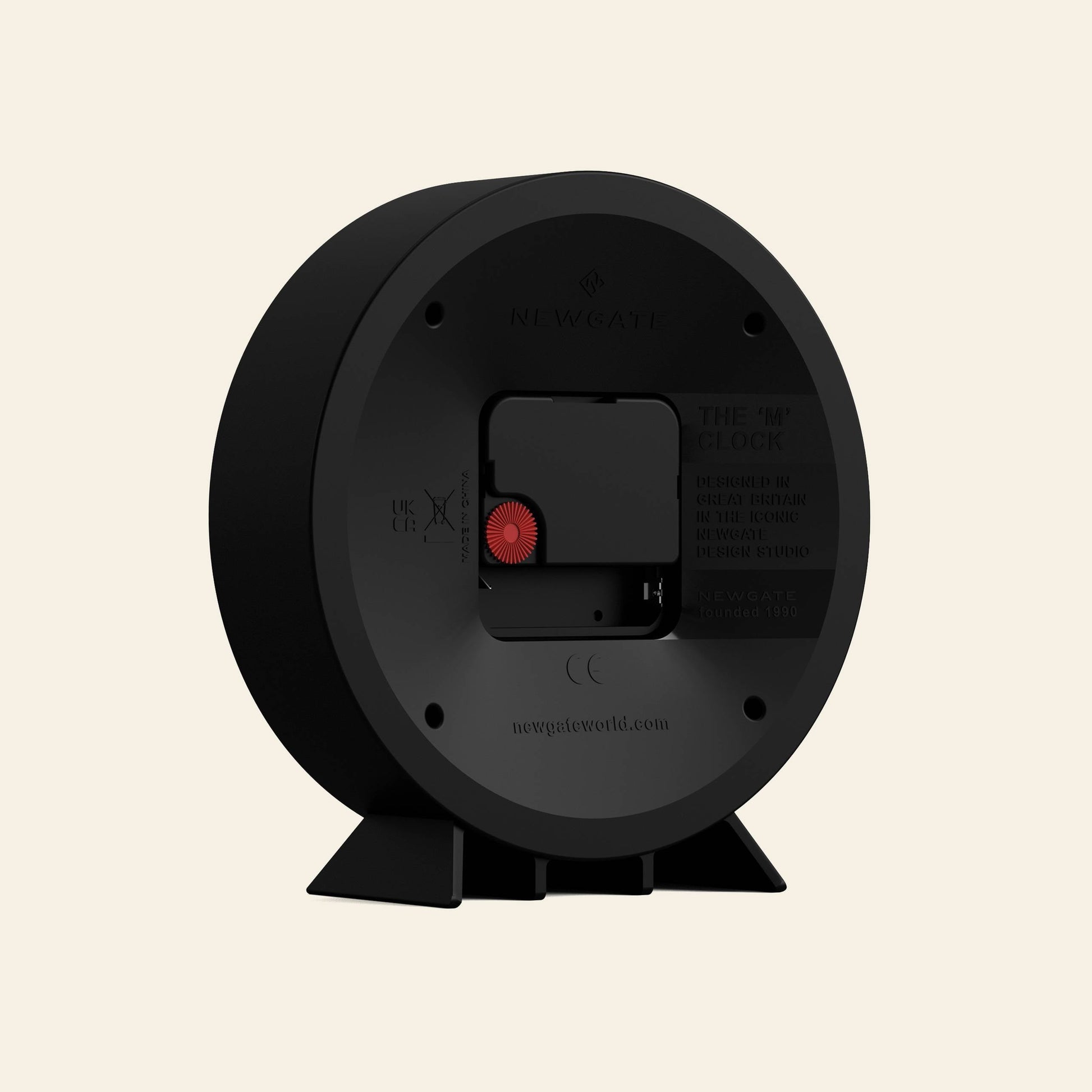 The Newgate M Mantel Railway Clock by Newgate, in black with a circular design, is shown from the back displaying its battery compartment, red adjustment dial, and product info. It stands on two angled feet against a light beige background.