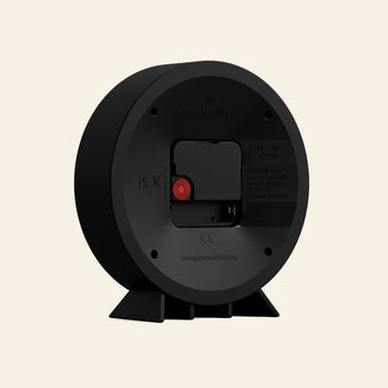 The Newgate M Mantel Railway Clock by Newgate, in black with a circular design, is shown from the back displaying its battery compartment, red adjustment dial, and product info. It stands on two angled feet against a light beige background.