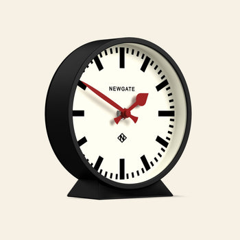 The Newgate M Mantel Railway Clock is a black, round analog mantel clock with bold black hour markers, two red hands set at 10:10 on a beige face, and NEWGATE branding—blending classic station style with modern minimalist design.