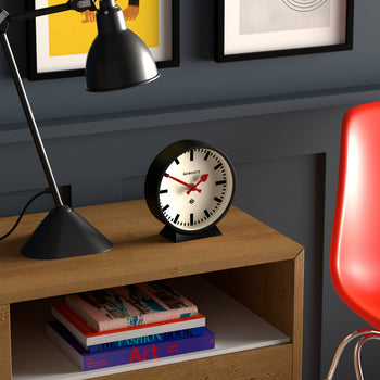 A modern desk with a black lamp, the Newgate M Mantel Railway Clock by Newgate, and a stack of colorful books beside a red chair. Framed artwork decorates the dark blue wall in the background.