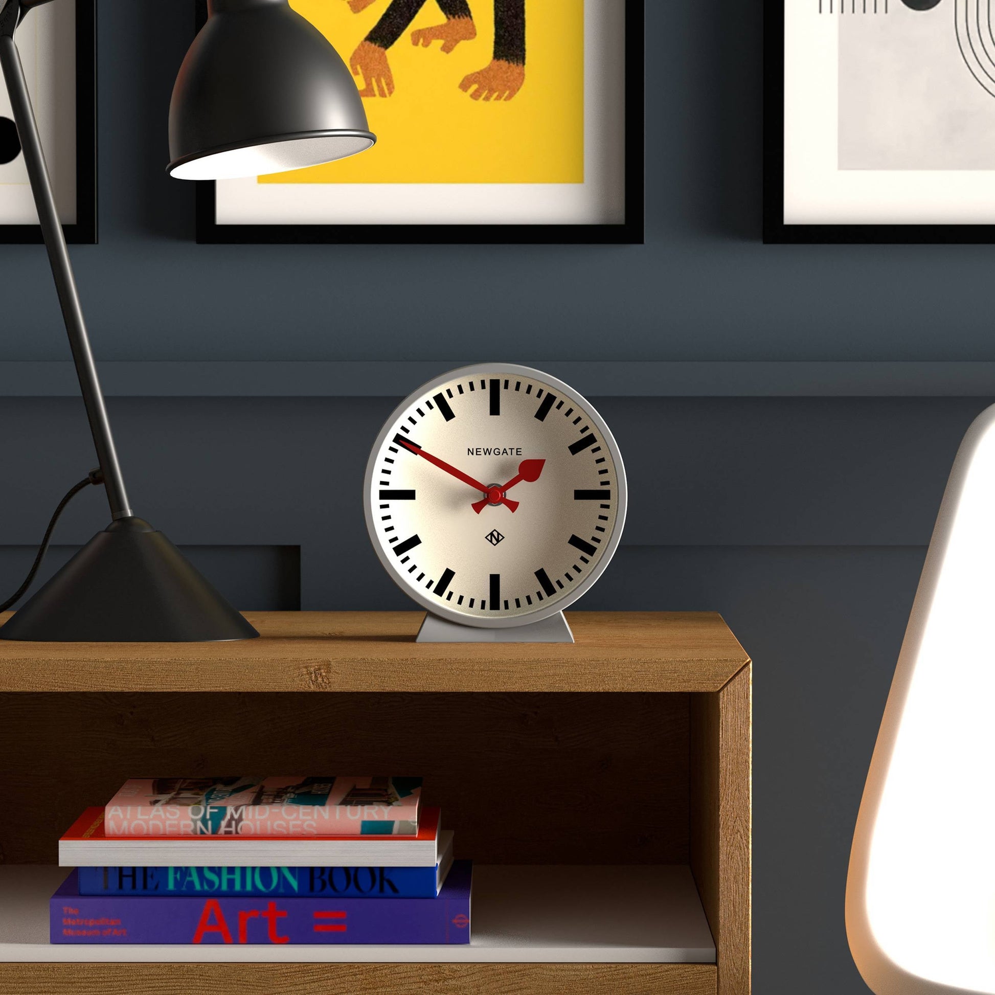 The Newgate M Mantel Railway Clock by Newgate, featuring red hands, is displayed on a wooden shelf beside a table lamp, with books and framed art set against a dark blue wall.